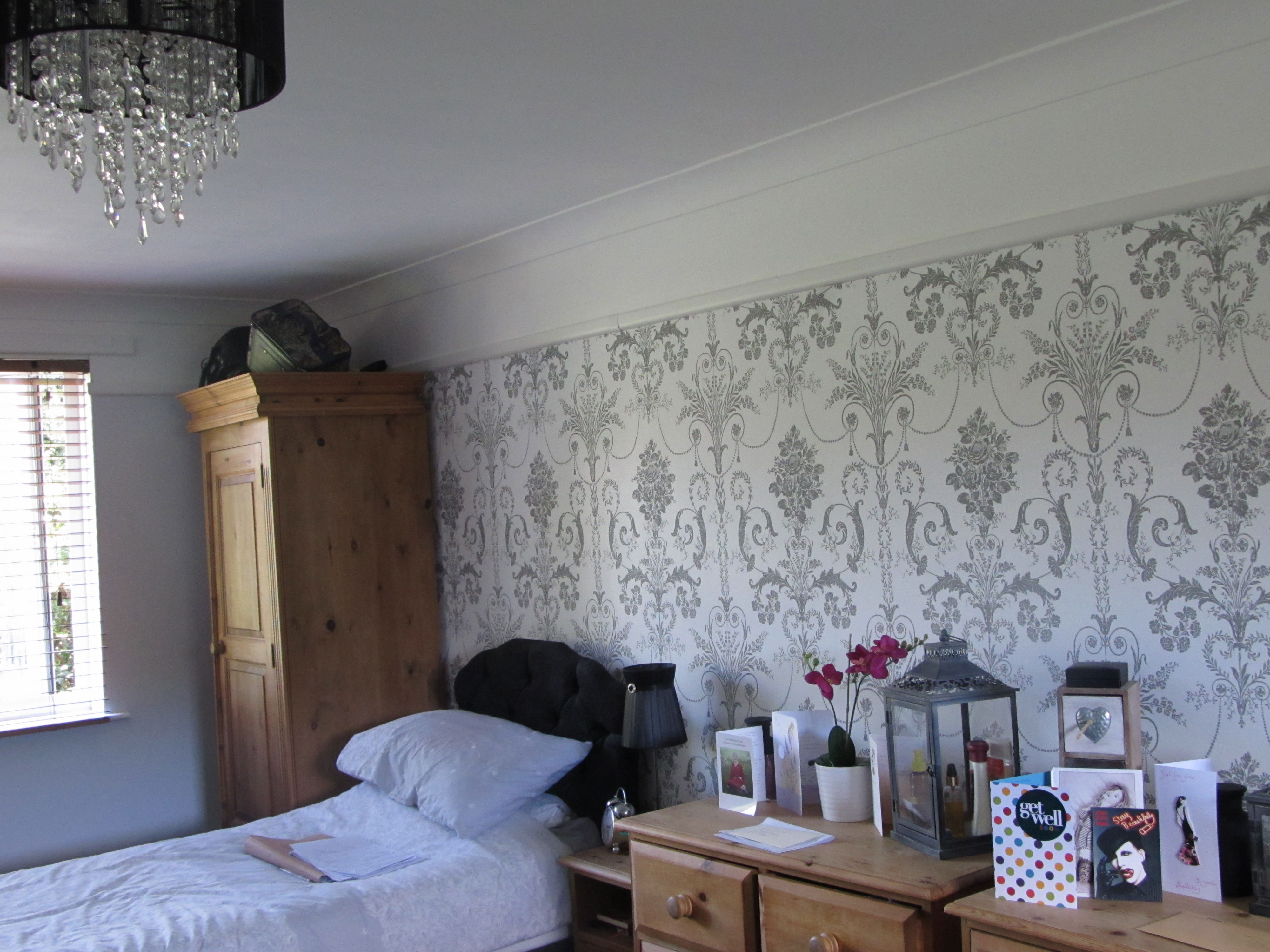 A photo of a bedroom at Allington House. There is an oak wardrobe in the left corner of the room, next to the single bed. There is a chest of drawers on the other side of the bed. The wallpaper is white and grey with a repeating baroque pattern