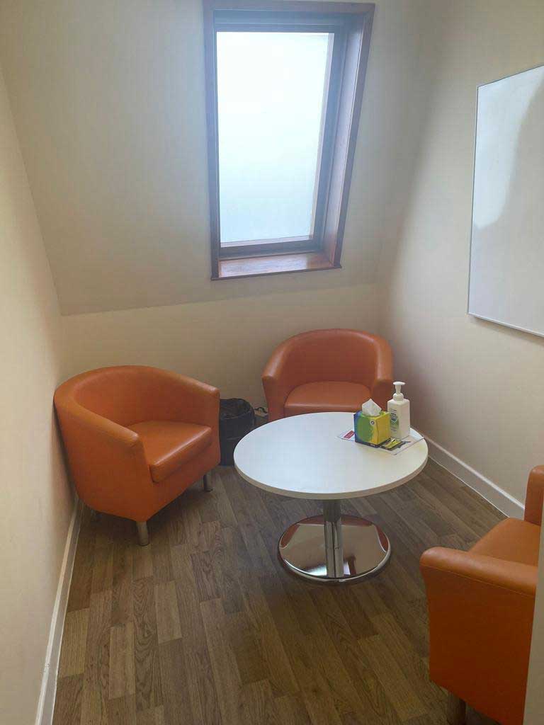 A small room with white walls, three orange tub chairs, a coffee table and a whiteboard