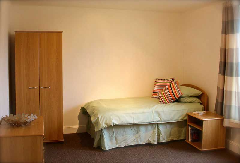 A bedroom with a single bed, a single wardrobe, bedside table and checked curtains