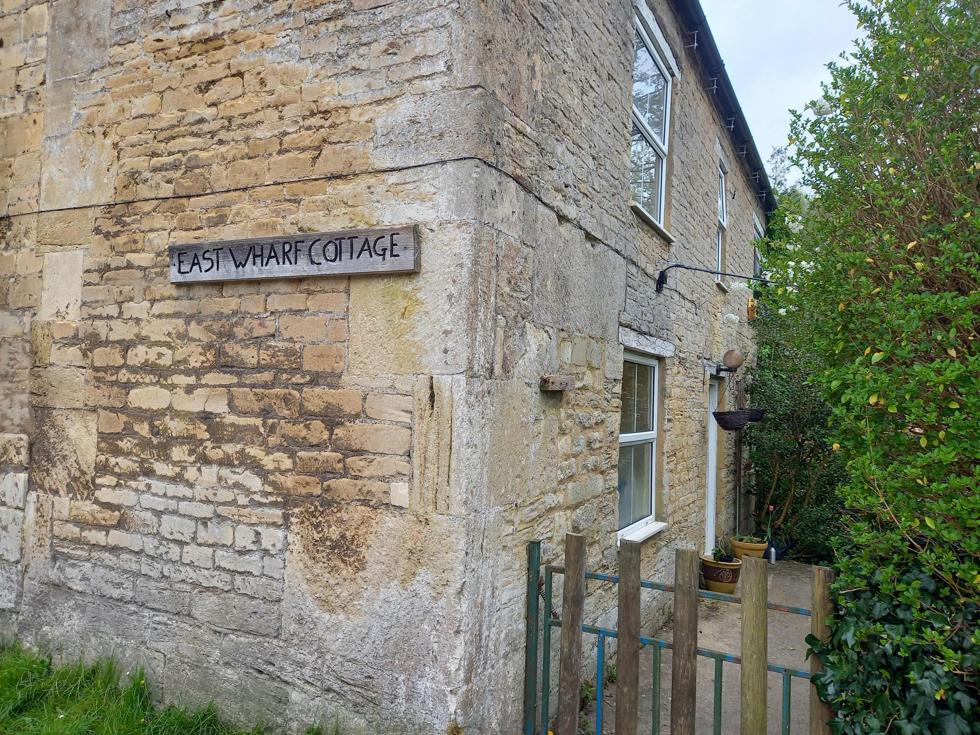 A close up of the corner of an old cottage. There is an old piece drift with the words 'East Wharf Cottage' engraved onto it. There is a small gate and a path leading to a door on the right hand side of the building, which is lined with green bushes.