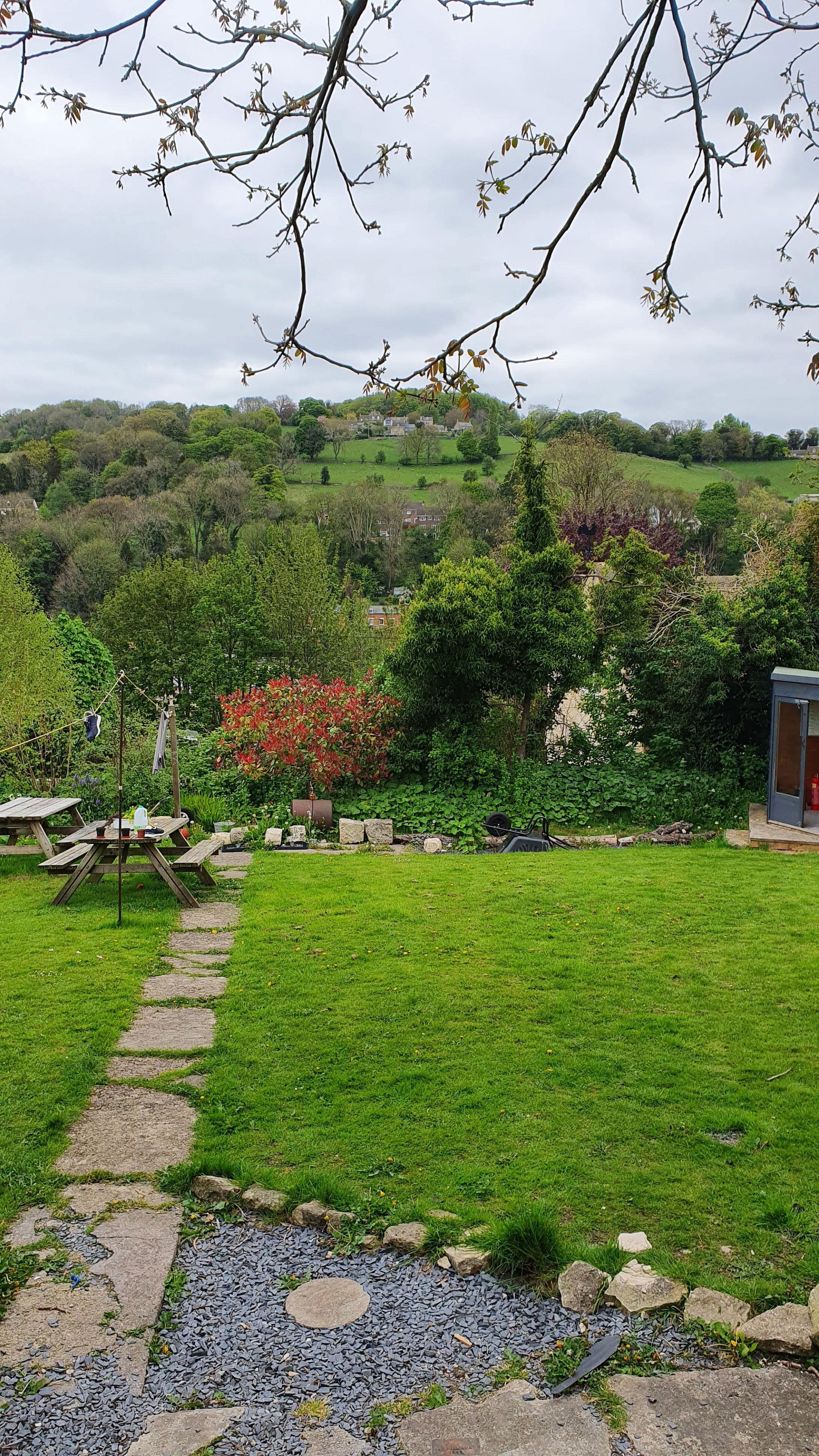 A photo of a garden overlooking rolling hills and fields. There is a small shed just visible at the bottom of the garden in the top right corner. A stone path leads to the bottom of the garden where a washing line is strung up between wooden poles. Two wooden picnic benches are also by the washing line.