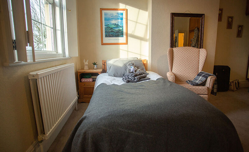A photo of a bedroom. There is a single bed in the middle of the image. On the wall to the left of the bed is a radiator and a bedside table. Above the radiator is a window with shutters. An arm chair sits to the right of the bed in front of a big mirror.