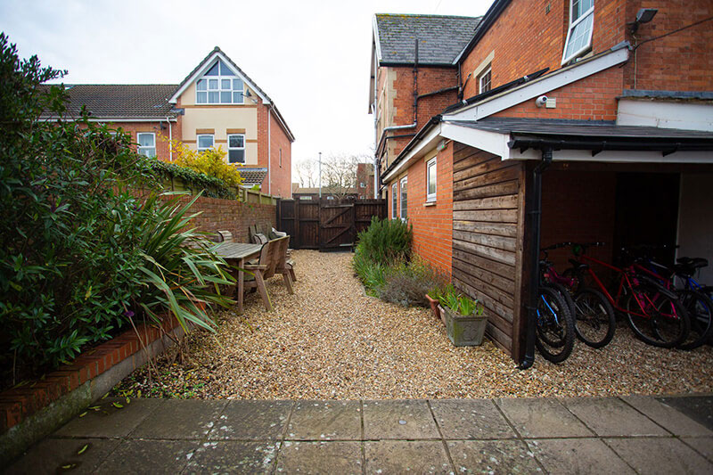 A photo of a garden. There is a bike shed on the right and a patio at the front of the image before it changes to gravel for the rest of the garden. There is a wooden table and chairs in the corner and plenty of plants lining the edges.