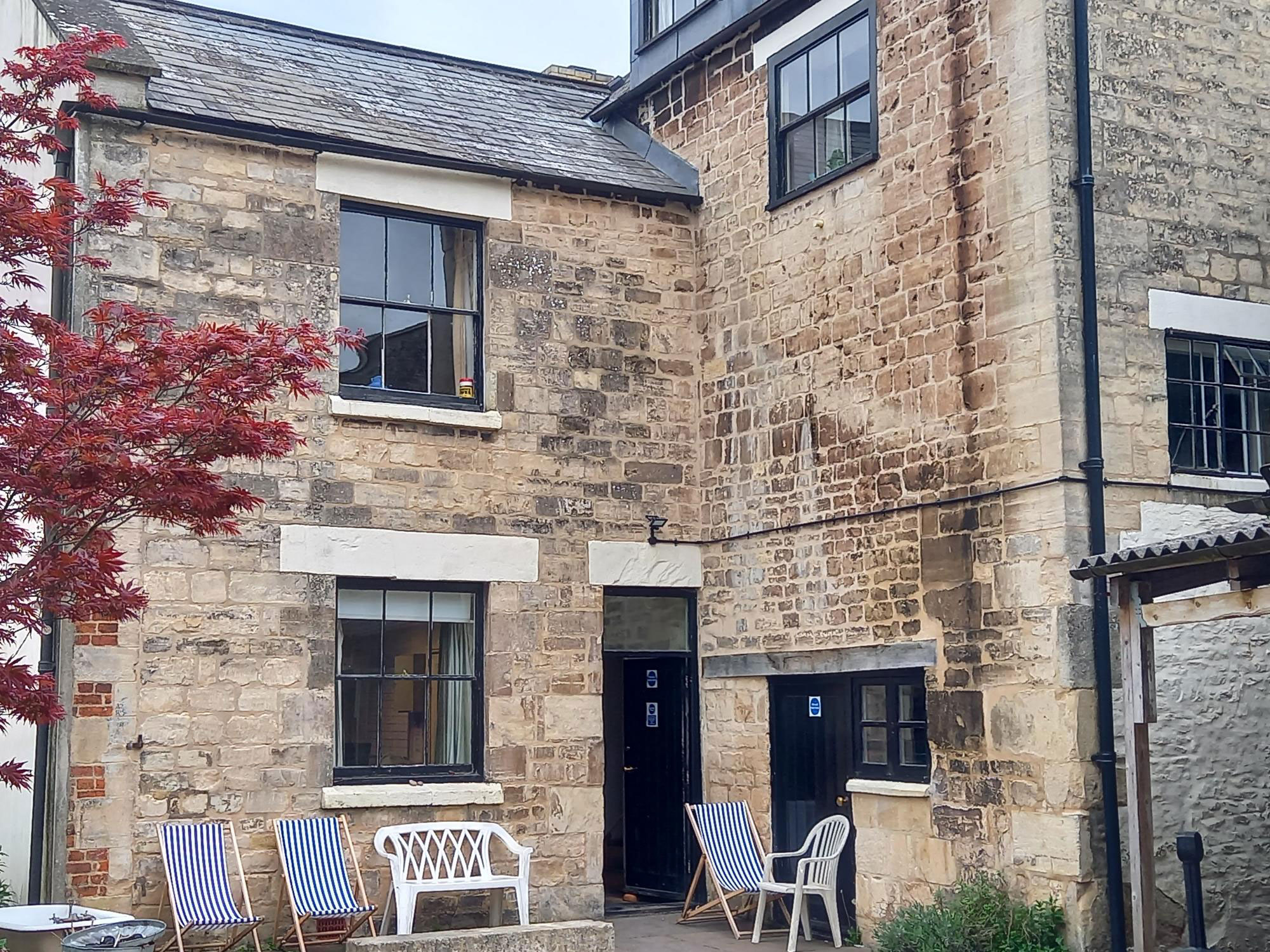 A photo of the courtyard of a house. There are a few windows dotted around the building and a door leading from the house to the courtyard. There are 3 blue and white striped deck chairs in the garden along with a plastic garden chair and two person plastic garden bench.