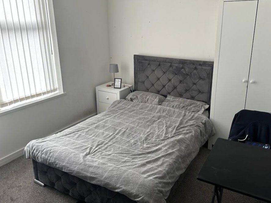 A photo of a bedroom - there is a grey double bed next to a window in a room with white walls. Next to the bed is a bedside table and a chair