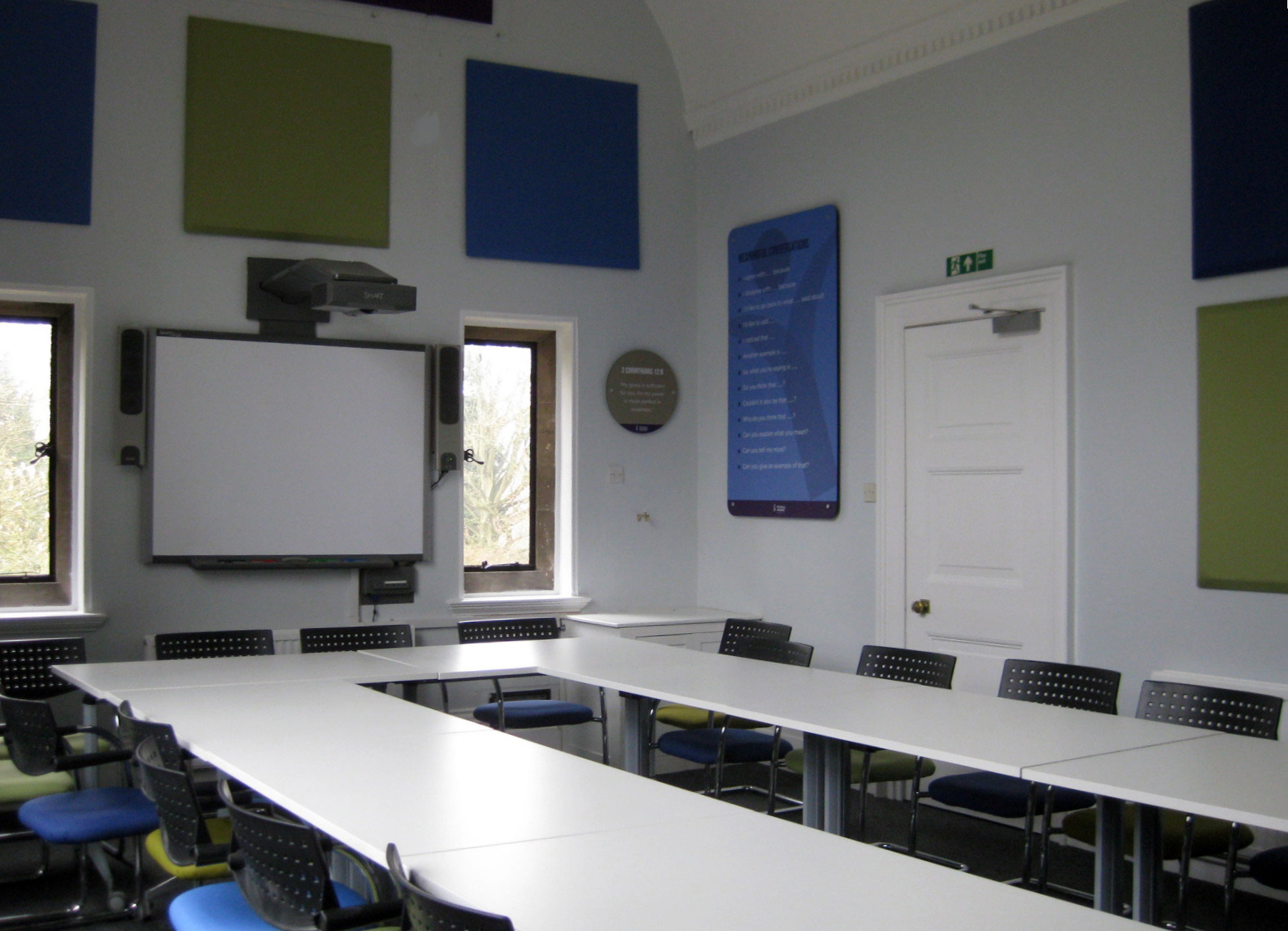 Yeldall Manor - group room. Tables form a U shape with chairs surrounding them. There is a whiteboard, windows and large colourful panels on the walls