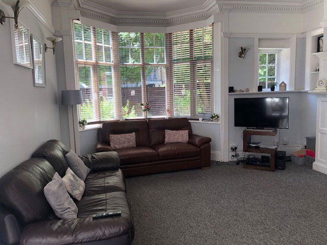 A photo of a communal area. There is a bay window along the back wall with venetian blinds. In front of the window is a two seater leather sofa, with another two seater leather sofa next to it. There is a TV in the corner of the room.