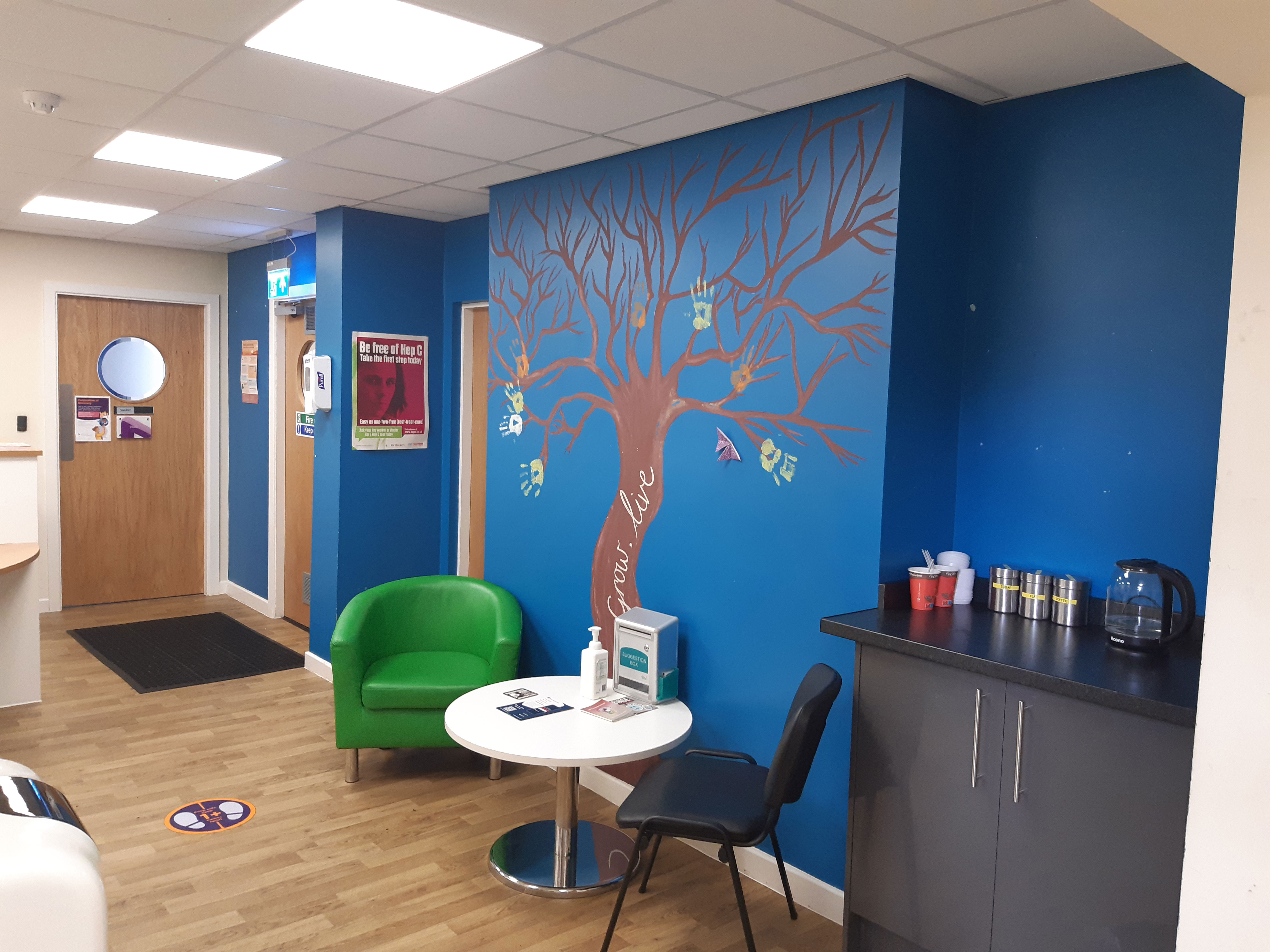 Macclesfield Recepton area - a room with a bright blue wall with a tree on it, green seating and white tables.