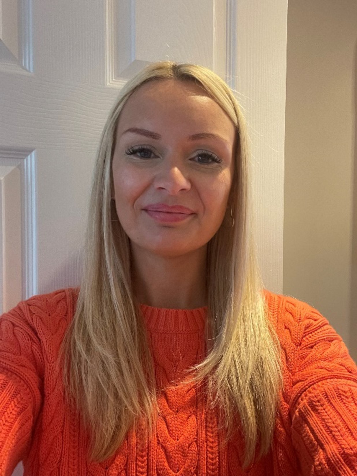 A photo of a blonde woman smiling at the camera. Her hair is down and rests below her shoulders. She is stood in front of a white door and wearing an orange jumper