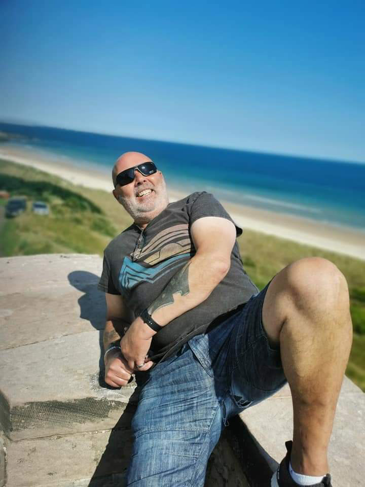 A photo of a man laying back, propped up on his arm. He is wearing a grey t-shirts and blue jean shorts. He is outside by the beach.