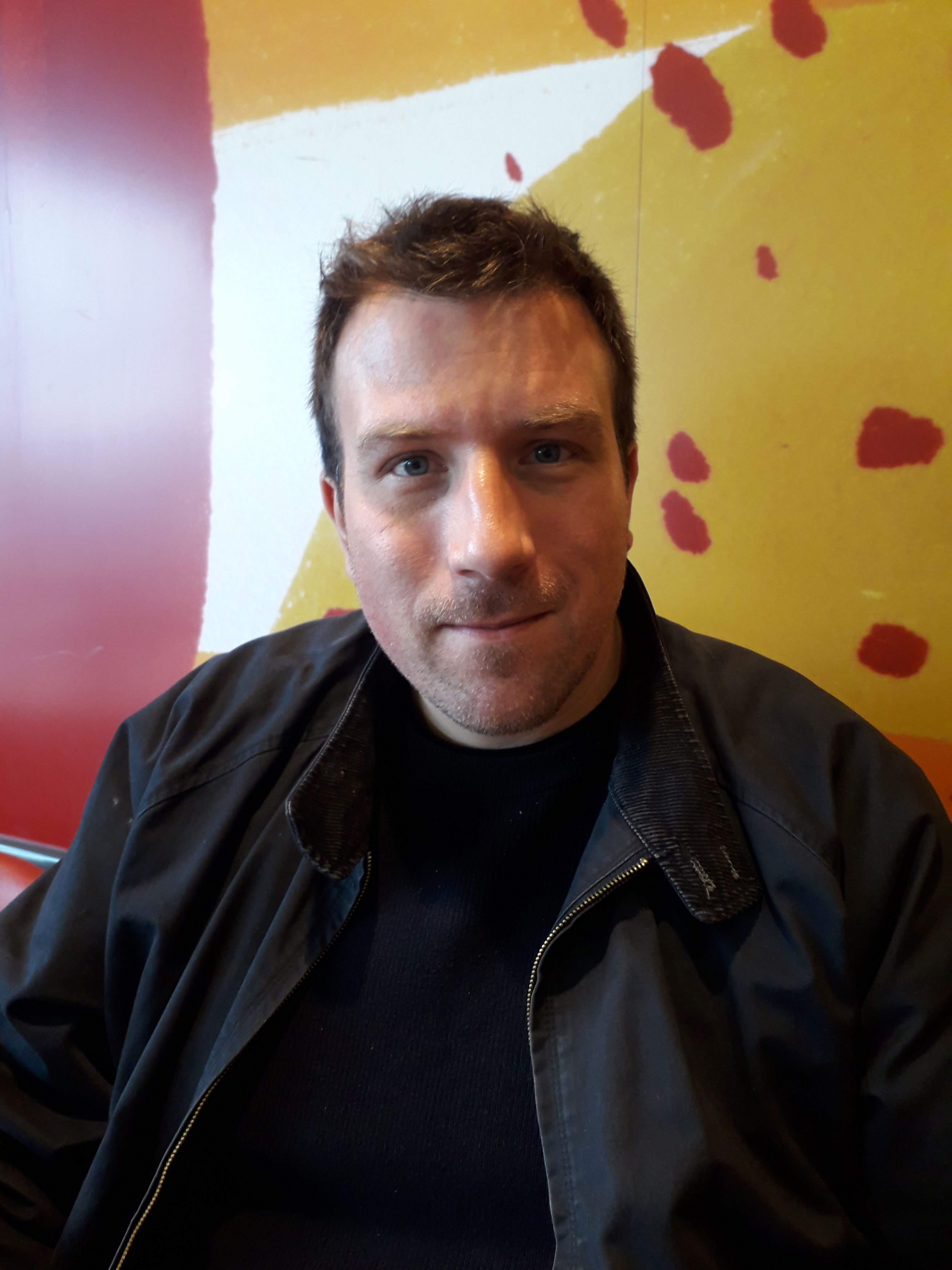 A photo of a man wearing a black t-shirt and black jacket. he is stood in front of a red and yellow wall