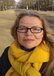 A photo of a women smiling at the camera. She is wearing black glasses, a yellow scarf and a black jacket. She has blond hair. 