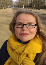 A photo of a women smiling at the camera. She is wearing black glasses, a yellow scarf and a black jacket. She has blond hair. 