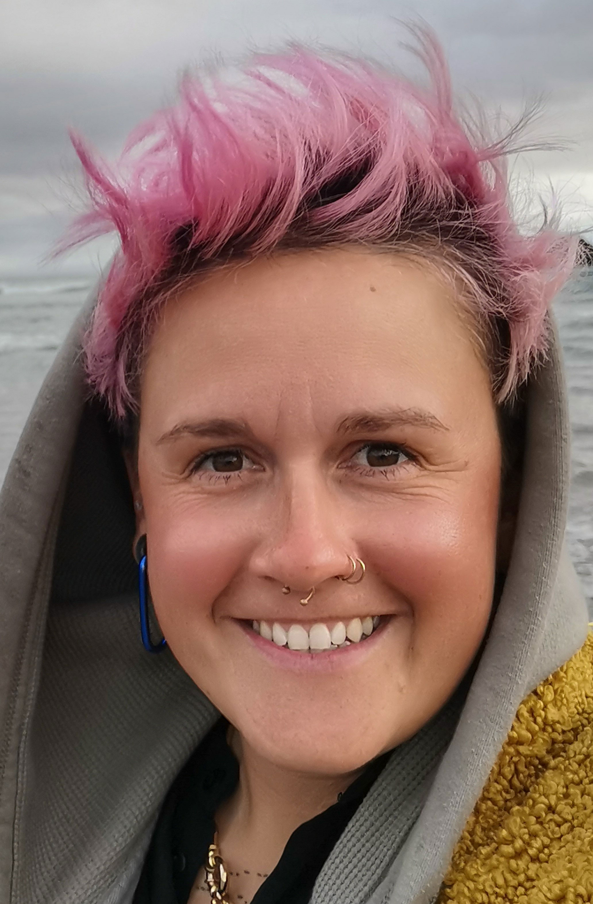 A photo of a woman smiling at the camera. She has bubblegum pink short hair which is sticking out from a grey hoodie