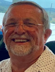 A photo of a man smiling at the camera. He has grey hair and a grey beard and is wearing glasses