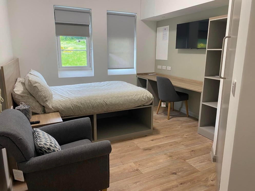 A single bed facing a desk with a TV above it. There is an armchair next to the bed.