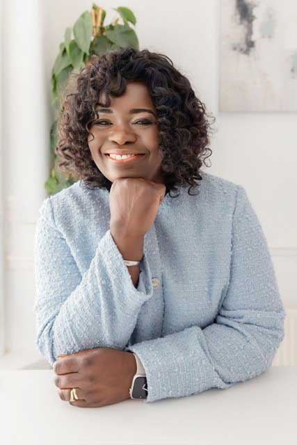 A professional headshot of a black woman. She is wearing a baby blue jumper and her curly hair is shoulder length