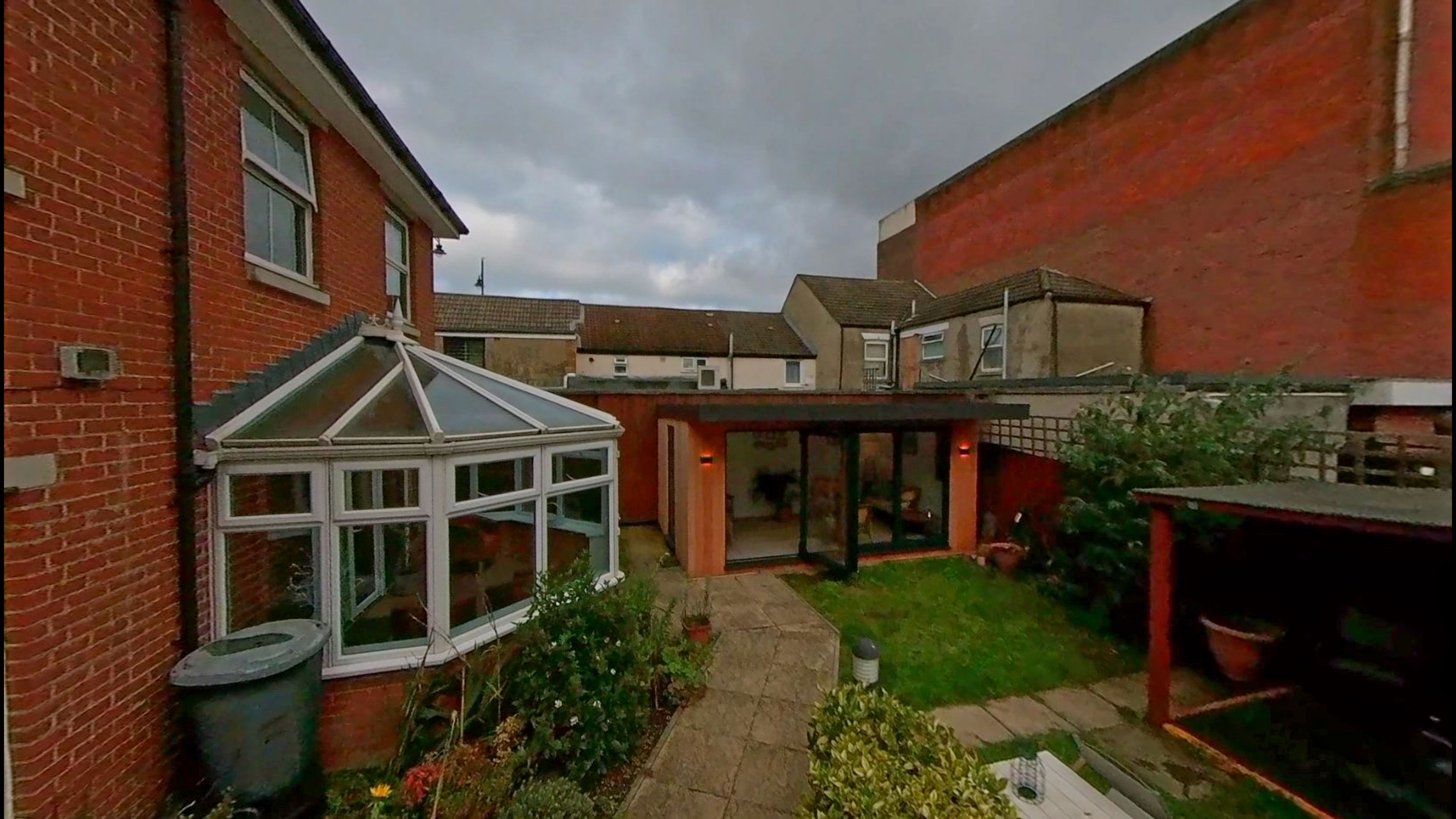 A garden with a conservatory and outbuilding. There is a small patch of grass with the rest of the space being pathed or planted with flowers