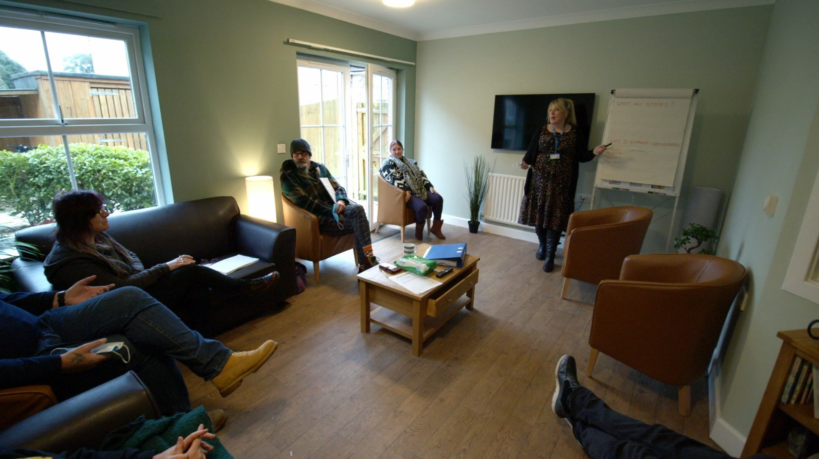 A room with leather sofas and chairs. A woman is stood at one end pointing at a flipchart while four other adults, are sat on the chairs looking at her.