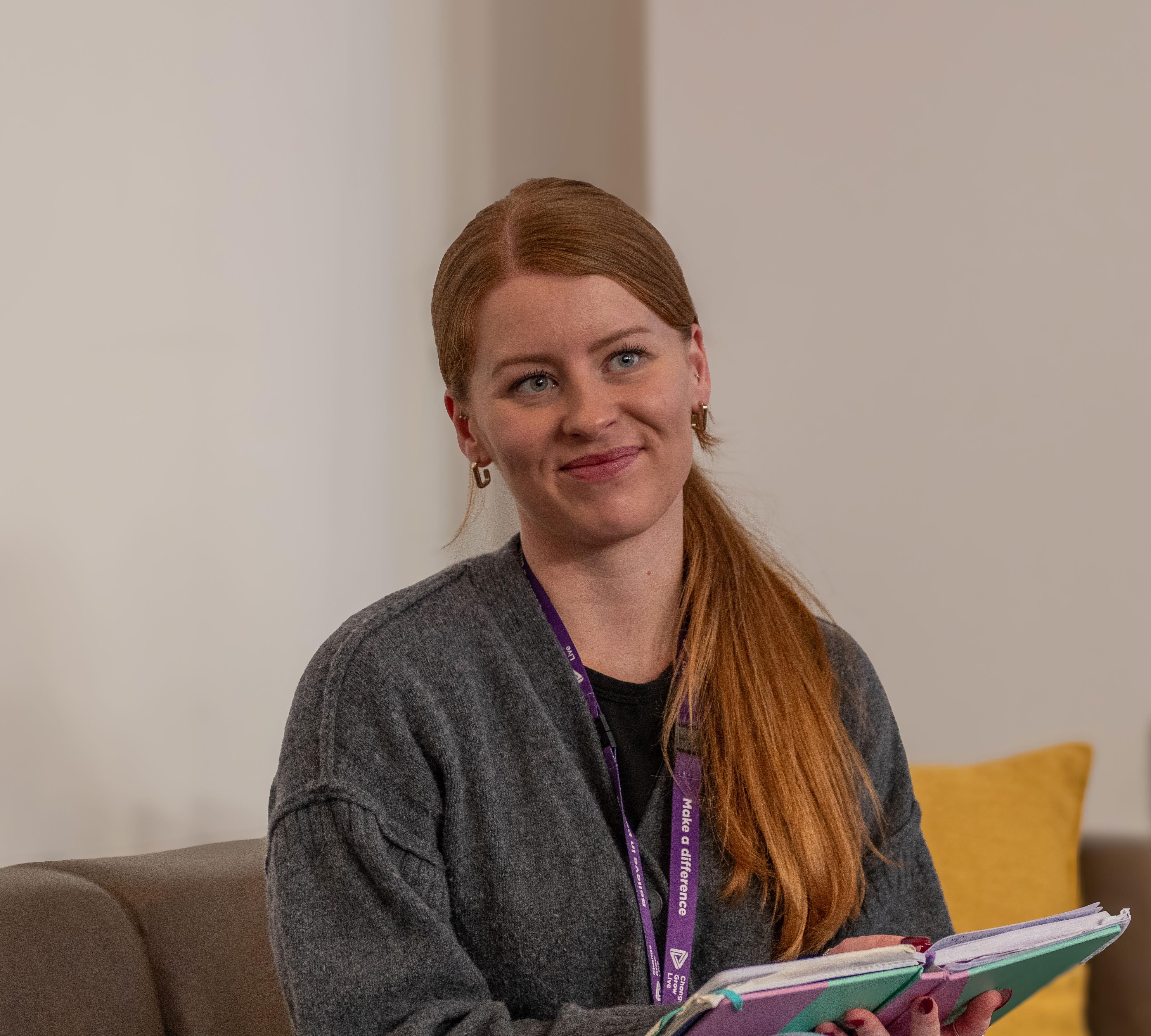 A young woman with ginger hair in a ponytail over her shoulder is sat on a sofa looking at someone off camera