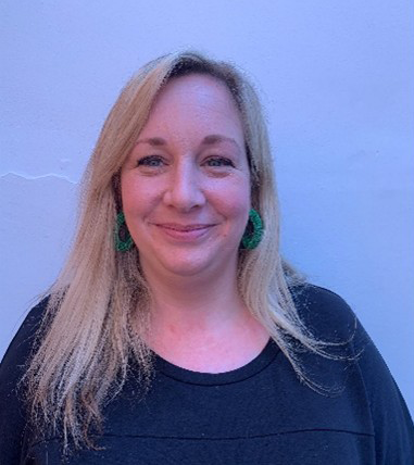 Mel has shoulder length blonde hair and is stood in front of a blue wall