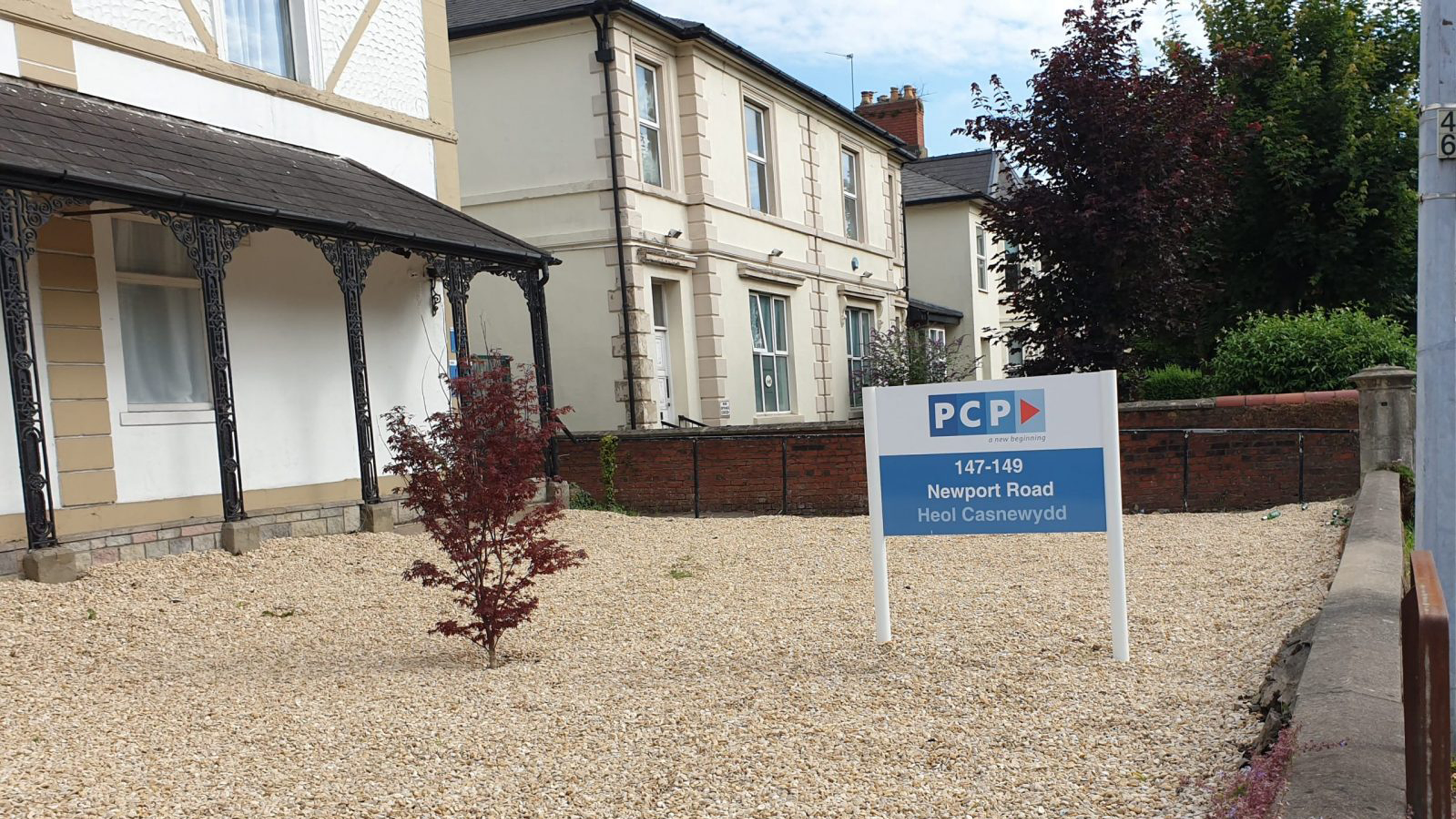 The outside of a building with a gravel drive. There is a sign in the drive advertising the establishment and a small tree with red leaves
