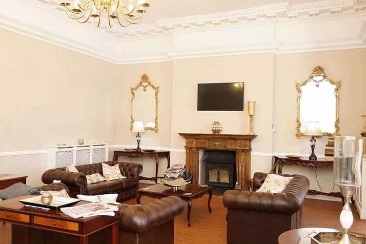 A lavish lounge area with brown leather sofas, a wooden fireplace with gold gilded mirrors hanging either side and a chandelier hanging from the ceiling