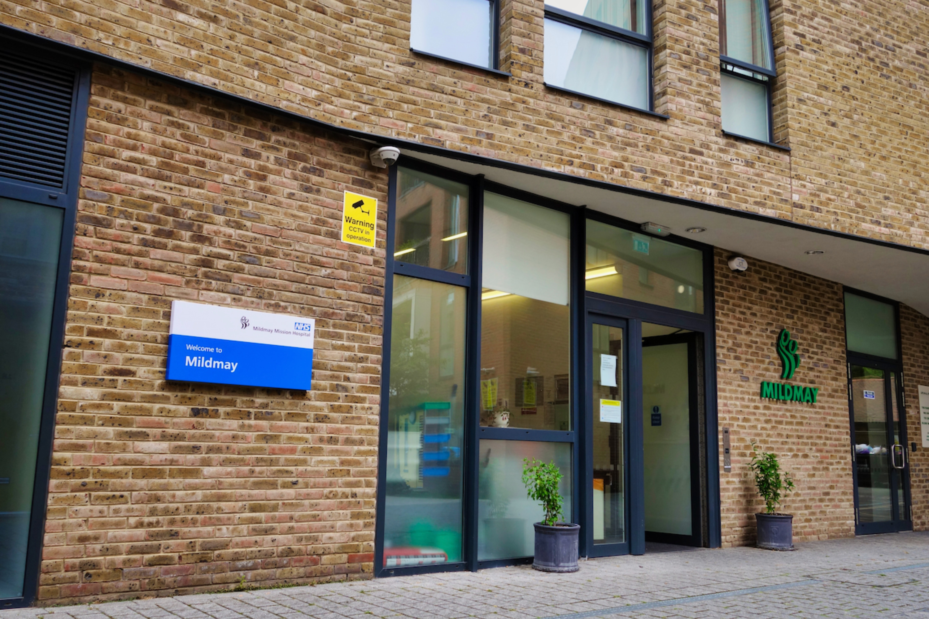 The front of a hospital with Brick wall and big glass double doors leading inside. A NHS sign is on the wall for Mildmay Hospital
