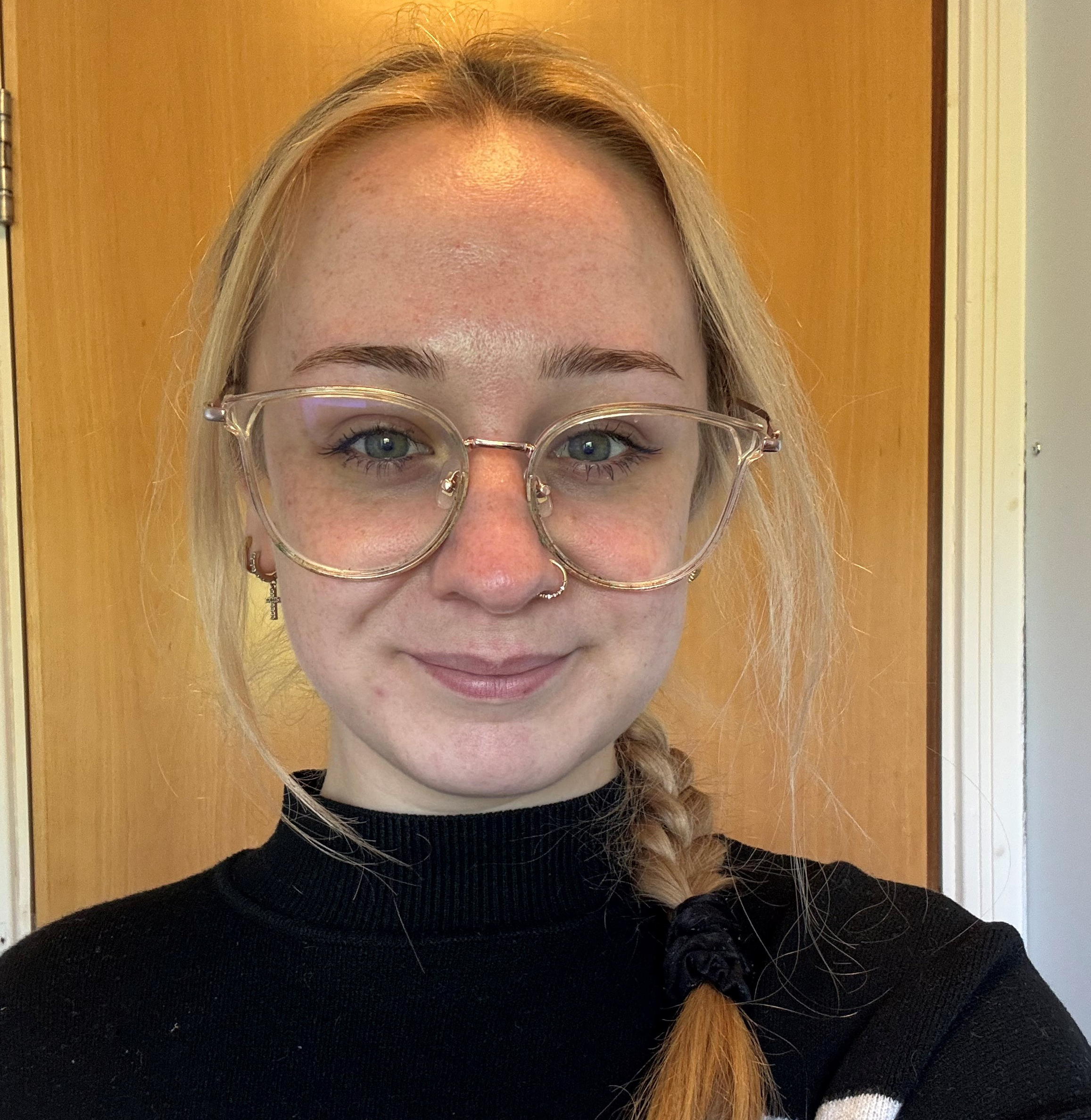 Becki is wearing glasses and a black turtleneck top. Her blonde hair is plaited and draped over her shoulder