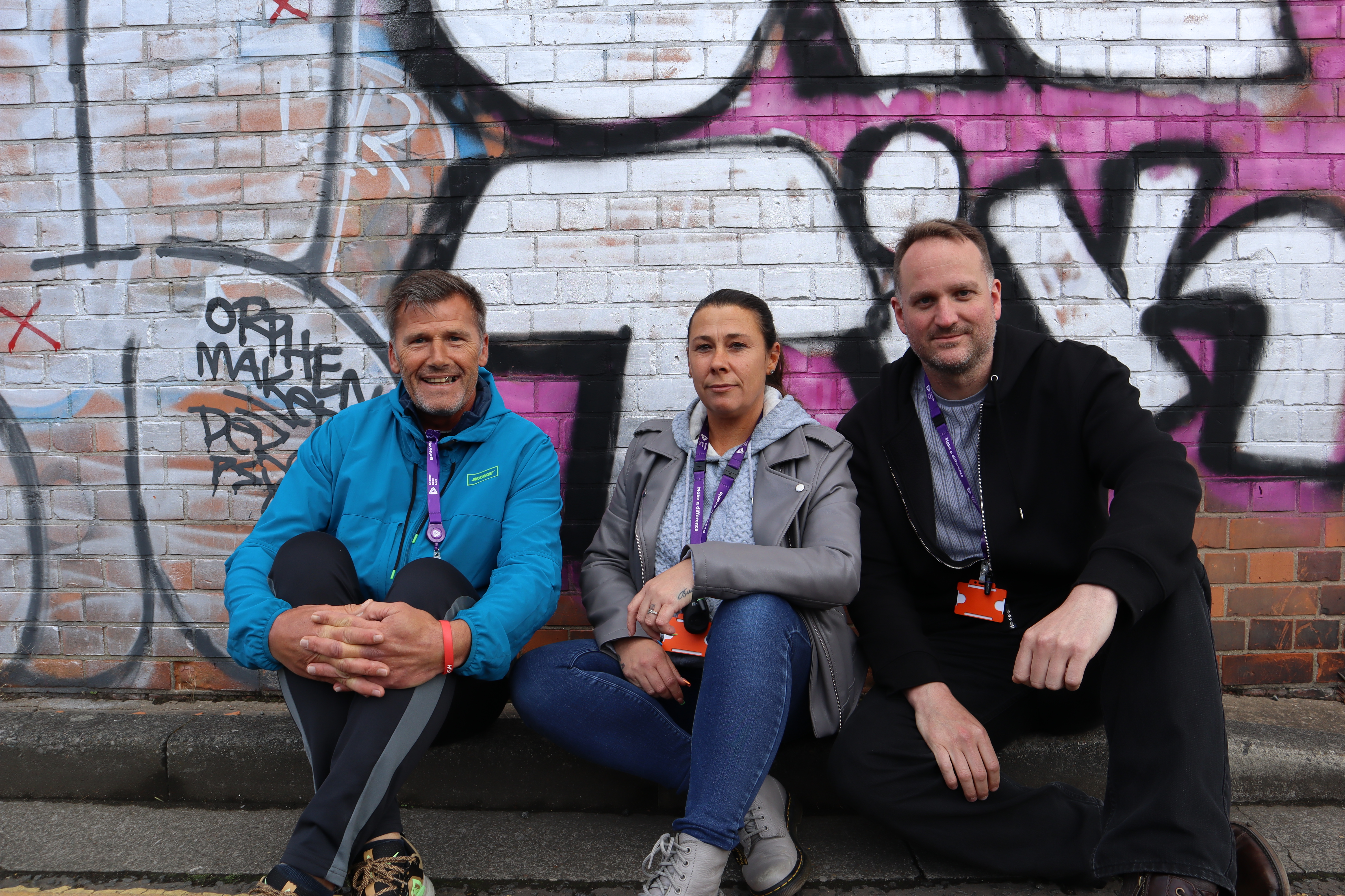 Three of the DWP project peer mentors sit in front of a colourful graffiti wall
