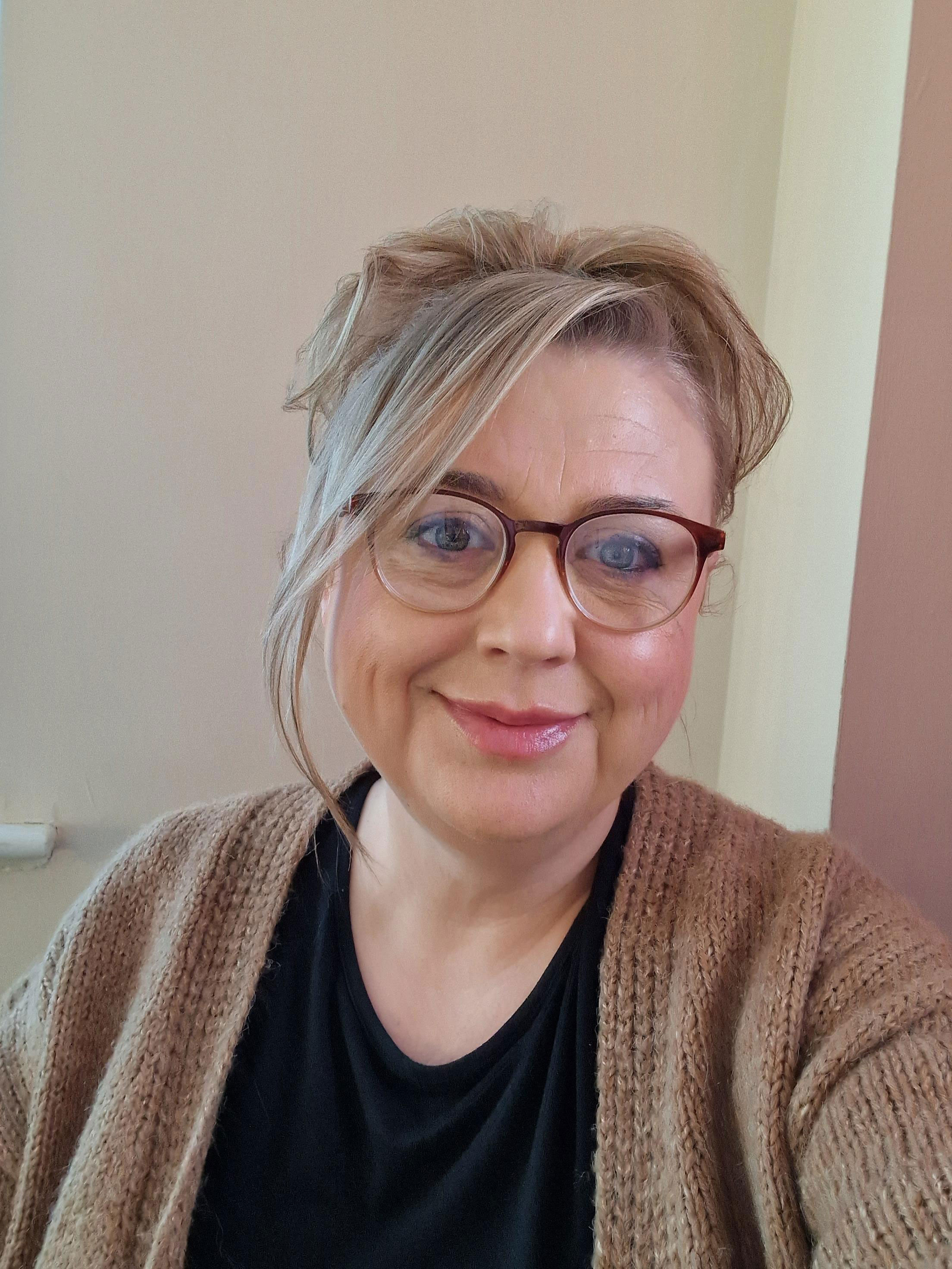 An older woman taking a selfie, her grey hair is pulled back and she is wearing a brown cardigan and glasses.