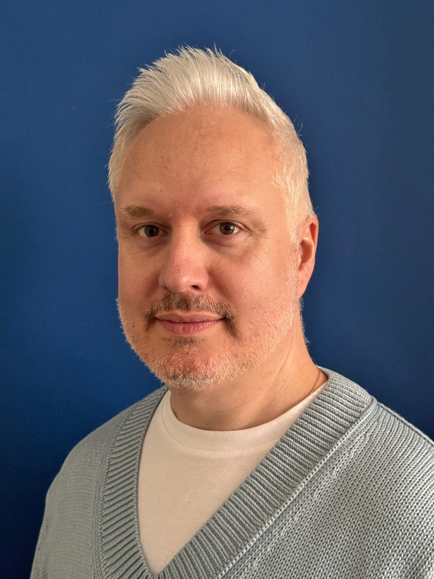 Lee has white hair and is wearing a blue v neck jumper over a white t-shirt