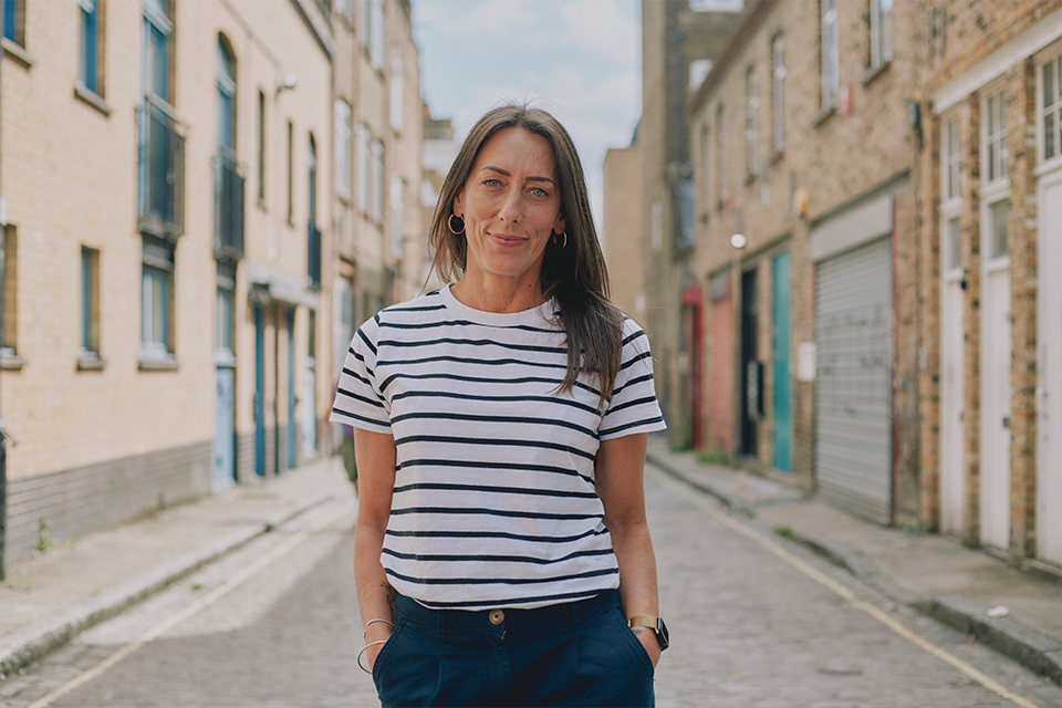 a woman in a blue and white striped t-shirt stands in an urban street setting smiling gently to camera