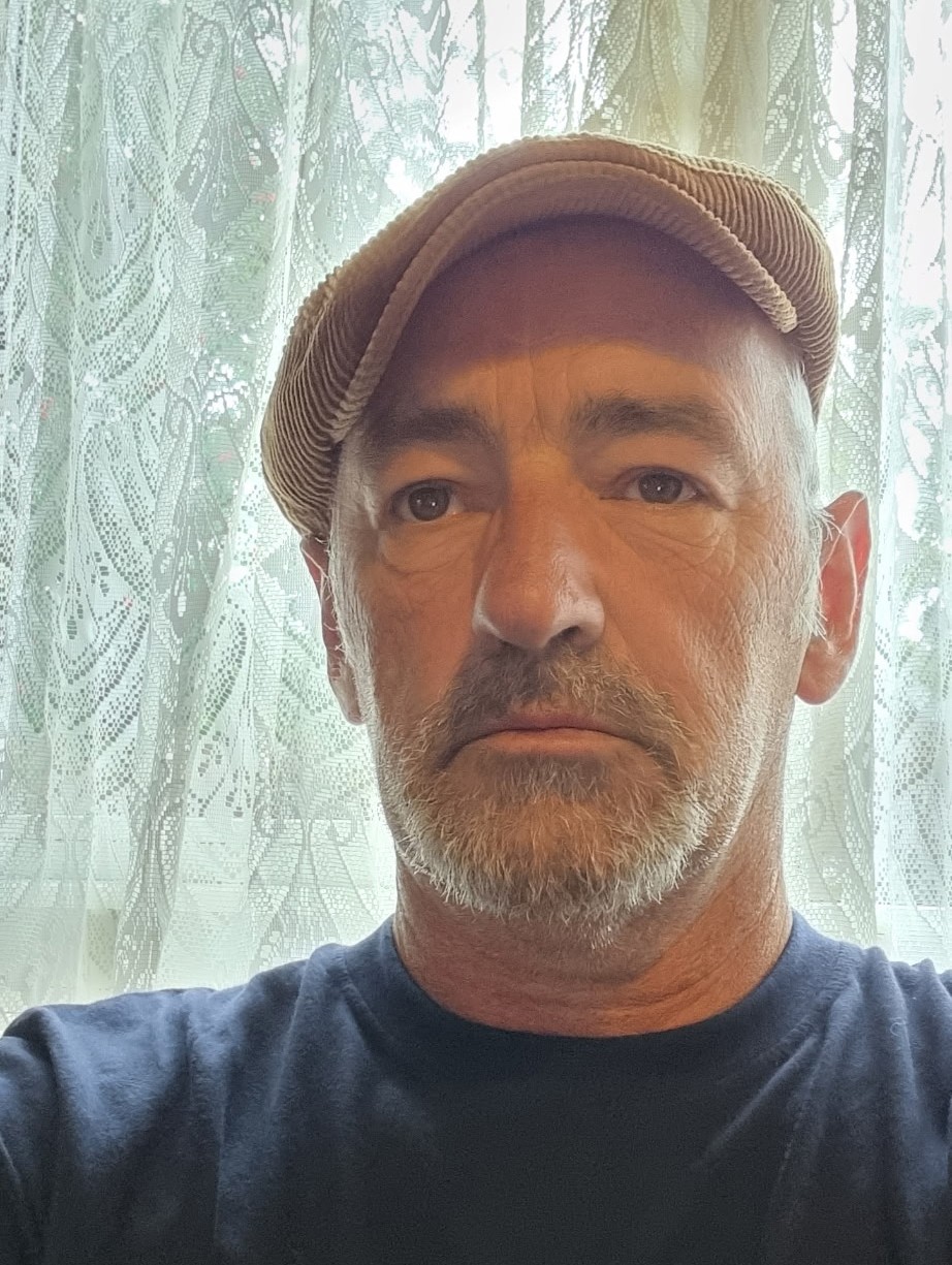 A middle aged man wearing a brown flat cap and a navy blue t-shirt. He has a slight beard and moustache.