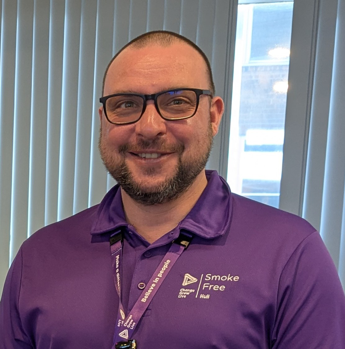A man wearing a purple polo shirt and glasses with a purple lanyard around his neck.