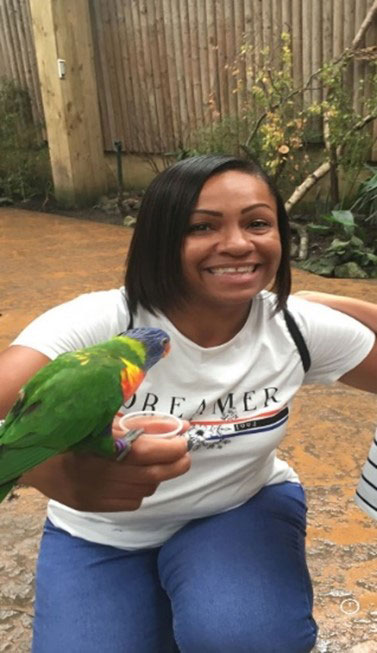 A photo of a woman with black hair. She is crouched to the ground, wearing a white t-shirt and blue jeans. She is holding a green parrot on her arm. 