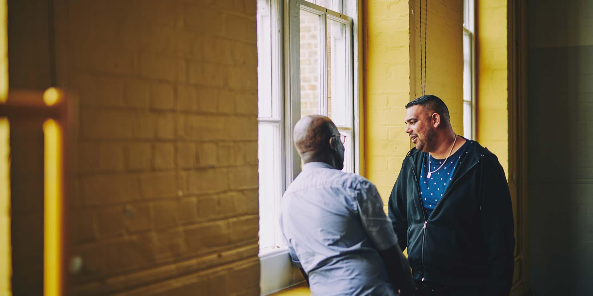 Two men are having a conversation by a window