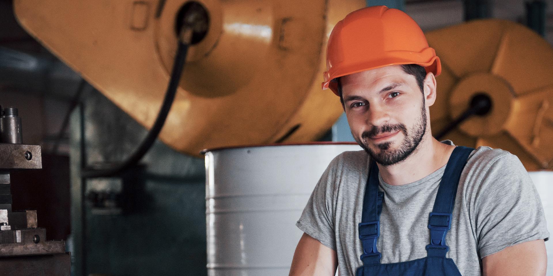 A man wearing an orange hard had and blue overalls stood in front of machinery