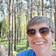 A woman with short cropped hair, wearing sunglasses and stood in a forest of trees.