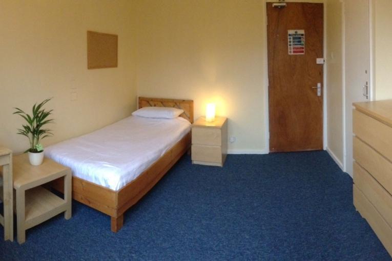 A photo of a bedroom , the bed is in the left hand corner, the door in the right, there is a chest of drawers in the front right corner of the photo. There are blue carpets and magnolia walls