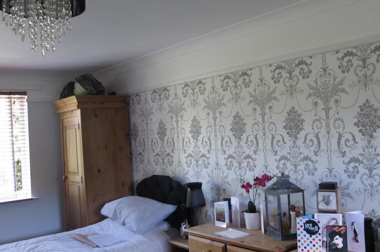 A photo of a bedroom at Allington House. There is an oak wardrobe in the left corner of the room, next to the single bed. There is a chest of drawers on the other side of the bed. The wallpaper is white and grey with a repeating baroque pattern
