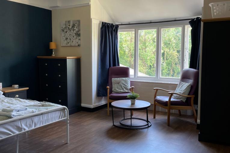 A photo of a bedroom. There is a big wide window along one wall with navy blue curtains. There are two leather arm chairs in front of the window with a circular coffee table. A chest of drawers sits in the corner next to a double bed. The walls are painted Navy and cream.