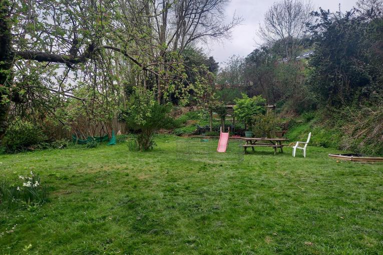 A photo of a big garden. There is plenty of open space and greenery, including grass and trees. There is a child's slide and a wooden picnic bench towards the back of the garden.