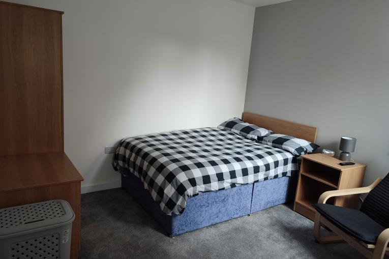 A photo of a bedroom. The room has grey walls and a grey carpet. There is a double bed in the corner of the room with a black and white gingham pattern on it. A bedside table sits next to the bed along with an arm chair. A pine wardrobe sits opposite the bed. 