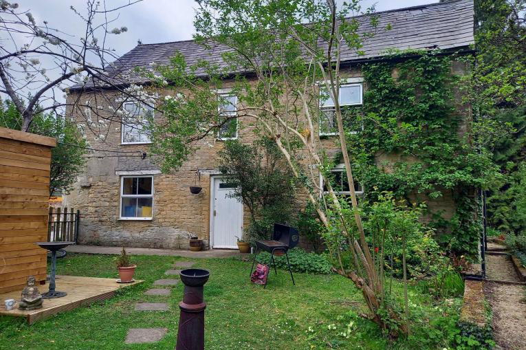 A photo of an old brick cottage with tiled rood. There is ivy growing up the side of with with 5 windows on the front of the building and a door. The garden has a stone path leading up to the door, with a wooden shed on the left. The shed is sitting on decking which also contains a bird bath, plant pot, and a small bhudda statue. On the right of the path is a bbq and a fire pit, along with trees and greenery. 