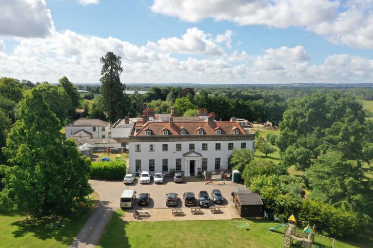 An aerial photographc of Kenworth House on a sunny day. A large white detached stately home with surrounding park. There are cars on a large drive