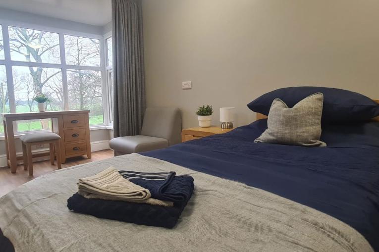 A photo of a double bed with navy blue sheets and a white blanket over the foot of the bed. There is a big window on the left side of the image. In front of the window is a small desk and stool. Next to the bed is a bedside table and a grey leather armchair.