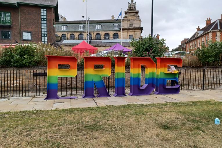 A photo taken of giant letters spelling out PRIDE. The letters are all painted in a rainbow from top to bottom. The Letters are stood infront of an iron fence in a park.