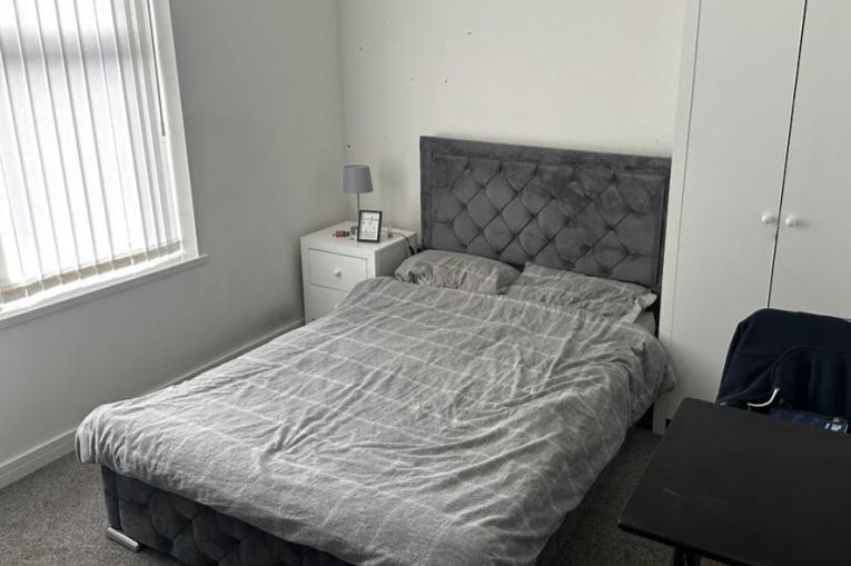 A photo of a bedroom - there is a grey double bed next to a window in a room with white walls. Next to the bed is a bedside table and a chair
