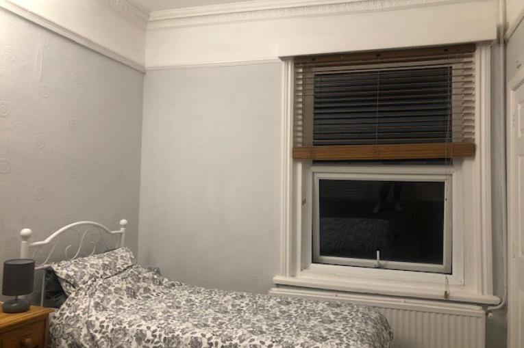 A photo of a bedroom. There is a single bed in the corner of the room with a window on the wall next to it with venetian blinds. Next to the bed is a small bedside table with a lamp on top. 